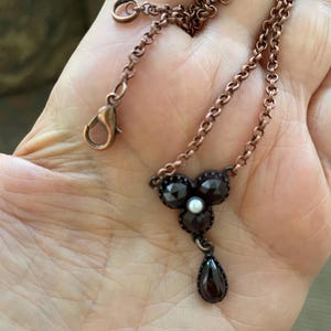 May include: A copper-toned necklace featuring a floral pendant with dark red gemstones and a small pearl center. A teardrop-shaped gemstone hangs below the floral design. The chain has a lobster clasp closure.