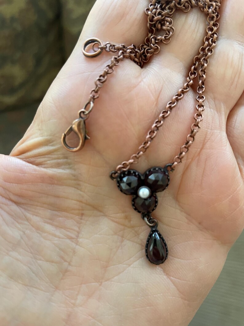 May include: A copper-toned necklace featuring a floral pendant with dark red gemstones and a small pearl center. A teardrop-shaped gemstone hangs below the floral design. The chain has a lobster clasp closure.