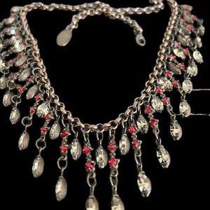 Może przedstawiać: Srebrny naszyjnik z łańcuszkiem i zwisającą frędzlą z przezroczystych i czerwonych kryształów.