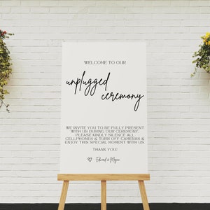 May include: A white sign on a wooden easel with the text "WELCOME TO OUR unplugged ceremony." The sign also includes a request to silence cellphones and enjoy the moment. The background is a white brick wall with hanging plants.