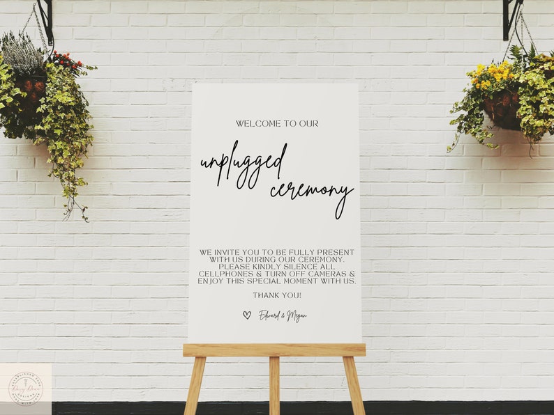 May include: A white sign on a wooden easel with the text "WELCOME TO OUR unplugged ceremony." The sign also includes a request to silence cellphones and enjoy the moment. The background is a white brick wall with hanging plants.