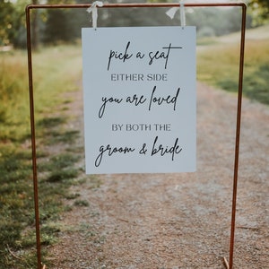 May include: A white sign with black script hangs from a copper-colored metal frame. The sign reads "pick a seat either side you are loved by both the groom & bride." The sign is outdoors on a gravel path.