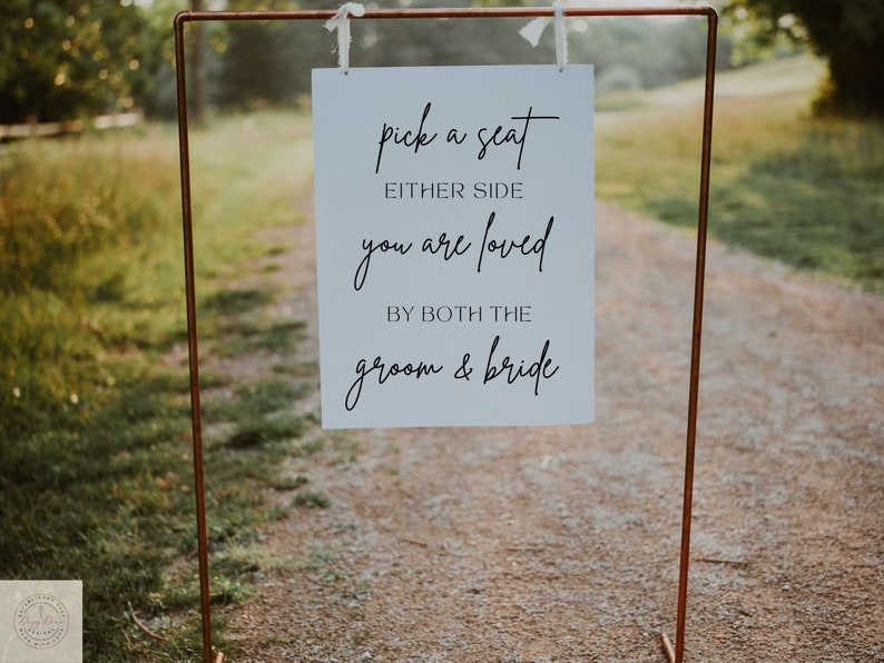 May include: A white sign with black script hangs from a copper-colored metal frame. The sign reads "pick a seat either side you are loved by both the groom & bride." The sign is outdoors on a gravel path.