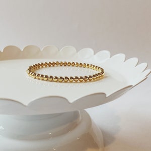 May include: A gold beaded bracelet on a white pedestal. The bracelet is made of small, round beads.