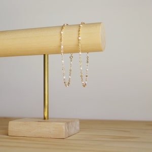 Puede incluir: Dos pulseras de cadena dorada con cuentas blancas colgando de un expositor de madera.