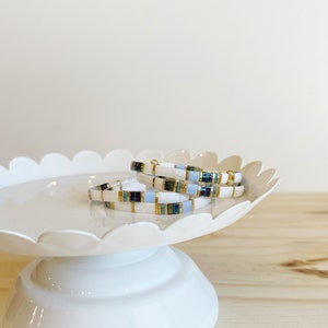 May include: Three white beaded bracelets with blue and gold accents stacked on a white cake stand.