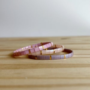 Könnte beinhalten: Drei übereinanderliegende Armbänder mit rosa, hellrosa und lila quadratischen Fliesen, die durch goldene Akzente getrennt sind. Die Armbänder liegen auf einer Holzoberfläche.