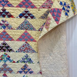 May include: A vintage quilt with a cream-colored background and colorful triangle patterns. The triangles feature various floral and geometric prints in red, blue, and white. The quilt is folded over, revealing a plain cream-colored backing.