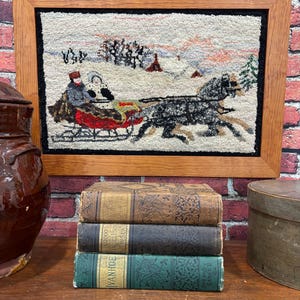 May include: A framed, vintage rug depicts a winter scene with a horse-drawn sleigh. The rug is in a wooden frame, above a stack of three antique books. The books are in various colors, including brown, black, and green, and are stacked on a wooden surface.