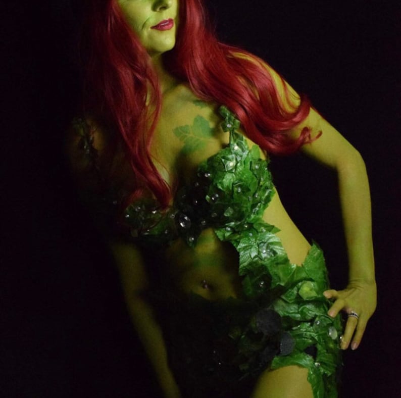 May include: A woman dressed as Poison Ivy, a DC Comics supervillain. She is wearing a green costume with leaves and vines, and has green makeup on her face and neck. Her hair is long and red.