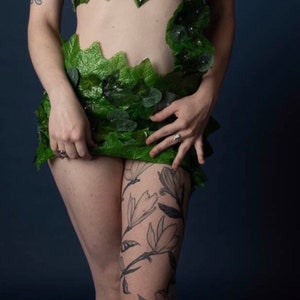 May include: A woman wearing a green leaf bra and skirt with a floral tattoo on her leg.