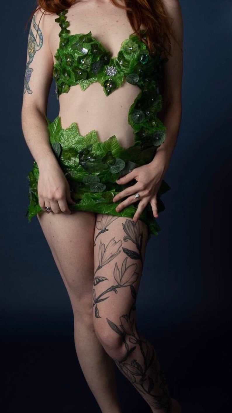 May include: A woman wearing a green leaf bra and skirt with a floral tattoo on her leg.