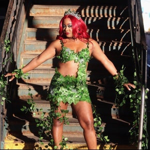 May include: A woman wearing a green leaf costume with a crown. She is standing on a set of stairs with ivy growing on the sides.