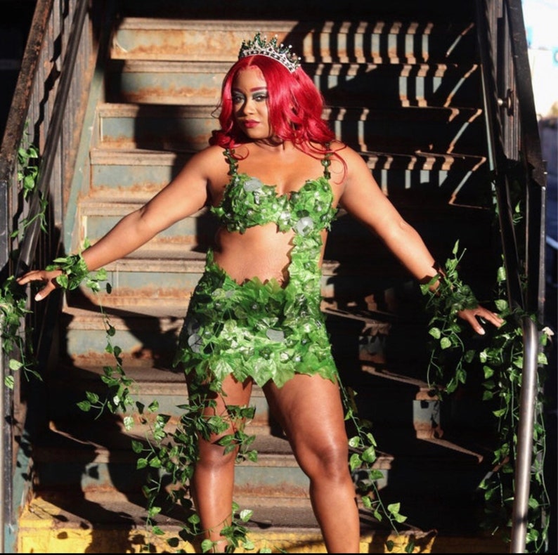 May include: A woman wearing a green leaf costume with a crown. She is standing on a set of stairs with ivy growing on the sides.