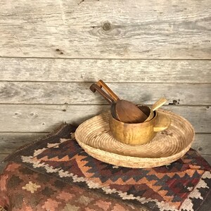 May include: A woven basket tray with a natural brown color, holding three wooden spoons and a wooden bowl. The tray is sitting on a colorful rug with a geometric pattern.
