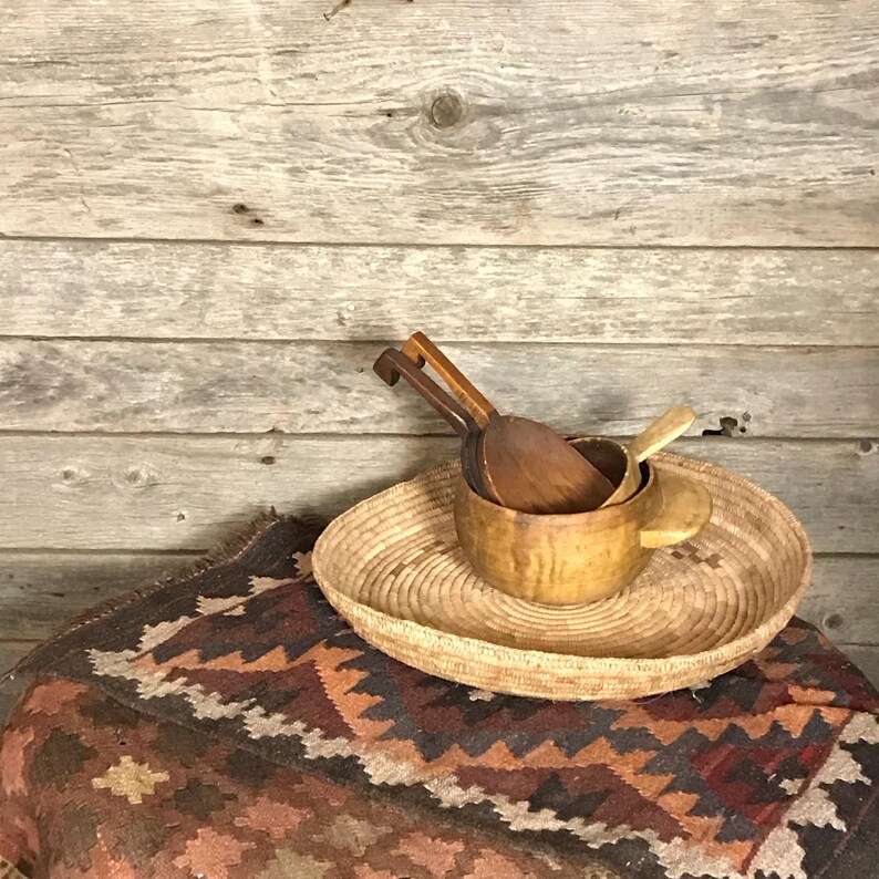 May include: A woven basket tray with a natural brown color, holding three wooden spoons and a wooden bowl. The tray is sitting on a colorful rug with a geometric pattern.