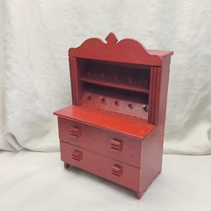 May include: A small, red wooden cabinet. It features two drawers and a hutch with two shelves and pegs. The cabinet is painted a solid red colour and has a decorative top. The cabinet's dimensions are approximately 30 cm in height.