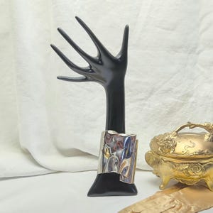 May include: A silver ring with a textured, folded design is displayed on a black hand-shaped jewellery stand. The ring has a shiny, reflective surface. A small, gold-coloured decorative box with a floral design is in the background.