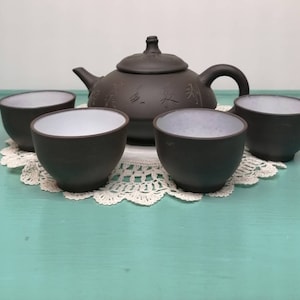 May include: A brown ceramic teapot with a lid and a matching set of four teacups. The teapot and cups have a white interior and are decorated with Chinese characters.