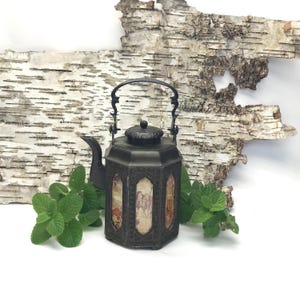 May include: An antique black metal teapot with a decorative handle and lid. The teapot has panels with images and is surrounded by fresh green mint leaves. The background features a piece of birch bark.