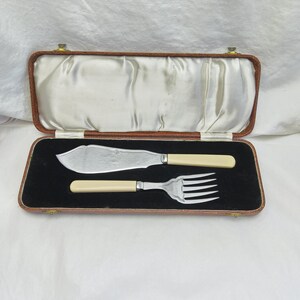 May include: A vintage butter knife and fork set with cream-coloured handles, displayed in a brown leatherette case with a black velvet interior. The knife has a wide, curved blade. The case is open, showing the silverware.