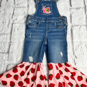 May include: Blue denim overalls with a pink and red strawberry print bell bottom. The overalls have a small pocket with a snail and strawberry patch. The denim has distressed details.