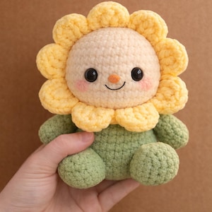Könnte beinhalten: Ein gehäkeltes Plüschtier in Form einer Sonnenblume mit cremefarbenem Gesicht, schwarzen Knopfaugen, einer kleinen orangen Nase und rosa Wangen. Die Blütenblätter sind hellgelb und der Körper ist salbeigrün mit abgerundeten Armen und Beinen.