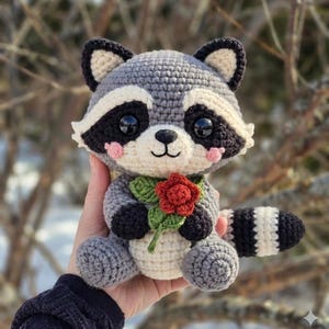 Könnte beinhalten: Ein handgefertigtes Häkel-Waschbär-Plüschtier in Grau, Weiß und Schwarz, das eine rote Rose hält. Der Waschbär hat einen gestreiften Schwanz und rosa Wangen. Das Plüschtier ist etwa 15 cm groß.