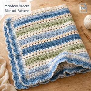 Könnte beinhalten: Eine gehäkelte Decke mit dem Muster "Meadow Breeze Blanket Pattern". Die Decke hat abwechselnde Streifen in Hellblau, Grün und Off-White mit einem blau gewellten Rand. Ein Wollknäuel und eine Häkelnadel sind sichtbar.