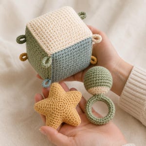 May include: A crochet sensory set featuring a cube with cream, sage green, and dusty blue panels, accented with small fabric loops. The set includes a five-pointed star in mustard yellow and a round rattle with a sage green ring and top.