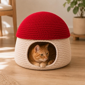 Könnte beinhalten: Ein kuppelförmiges Haustierbett aus Häkelarbeit mit einer leuchtend roten Oberseite und einer cremefarbenen Basis mit einer kreisförmigen Öffnung. Eine orangefarbene getigerte Katze ruht im Inneren der Struktur, die einem Pilzhut ähnelt.