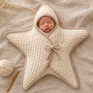 Peut inclure: Une gigoteuse pour bébé en crochet en forme d'étoile de couleur crème avec une capuche. Elle présente un motif de points texturés, une bordure marron clair sur les bords et un petit nœud de fermeture au centre. La forme est celle d'une étoile.