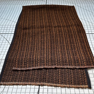 Puede incluir: Una toalla de cocina tejida de color marrón y naranja con un borde negro. La toalla está doblada por la mitad y tiene un patrón texturizado.