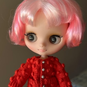 May include: A Blythe doll with a pink and white wig and large, expressive eyes. The doll is wearing a bright red, textured knit cardigan with silver buttons. The doll's skin is pale, and the overall aesthetic is whimsical and charming.