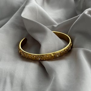 May include: Gold-coloured cuff bracelet with a celestial design. The bracelet features embossed suns, moons, and stars, with small, clear gemstones. The bracelet is set against a backdrop of soft, grey fabric.