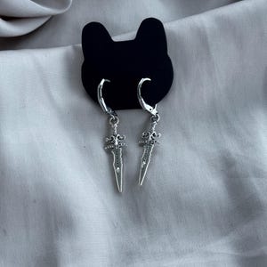 Könnte beinhalten: Silberfarbene Dolch-Ohrringe mit Klappbügelverschlüssen. Die Ohrringe werden auf einer schwarzen, katzenförmigen Ohrringkarte vor einem hellgrauen Stoffhintergrund präsentiert. Die Dolche haben detaillierte Griffe und spitze Klingen.