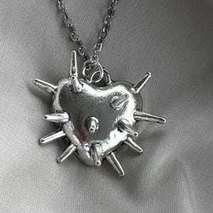 Könnte beinhalten: Ein herzförmiger Anhänger aus silberfarbenem Metall, der an einer passenden Gliederkette hängt. Der herzförmige Anhänger hat eine glatte, reflektierende Oberfläche, die mit mehreren hervorstehenden Stacheln an der Vorderseite und den Kanten verziert ist.
