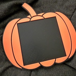 May include: Orange pumpkin-shaped picture frame with a black center. The frame has a black outline and a stem. The frame is made of a rubbery material. Perfect for Halloween or fall decor.