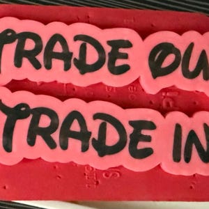 May include: A rectangular red silicone mold featuring two raised, scalloped-edge text sections in pink. The top section displays the black text TRADE OUT, and the bottom section displays the black text TRADE IN, both in a bold, rounded font.