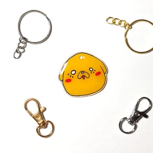 May include: A yellow cartoon dog charm with red cheeks and a black nose, attached to a silver key ring. There are three other key rings in the image, one silver and two gold.