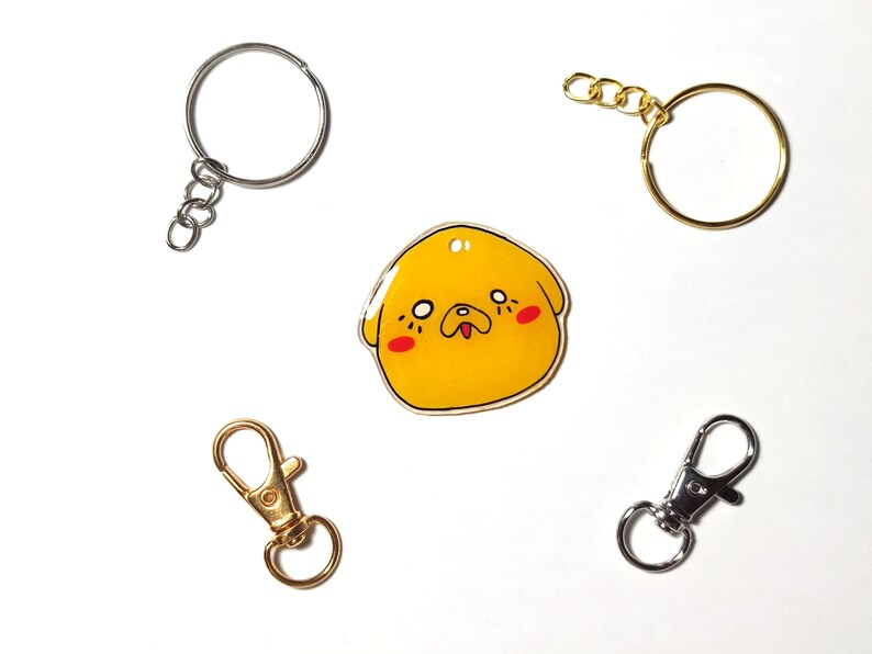 May include: A yellow cartoon dog charm with red cheeks and a black nose, attached to a silver key ring. There are three other key rings in the image, one silver and two gold.