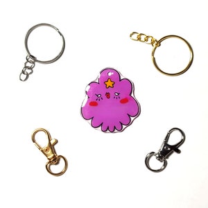 May include: A purple cartoon character keychain with a star on its head. The keychain is attached to a silver key ring. There are three other key rings in the image, two silver and one gold.