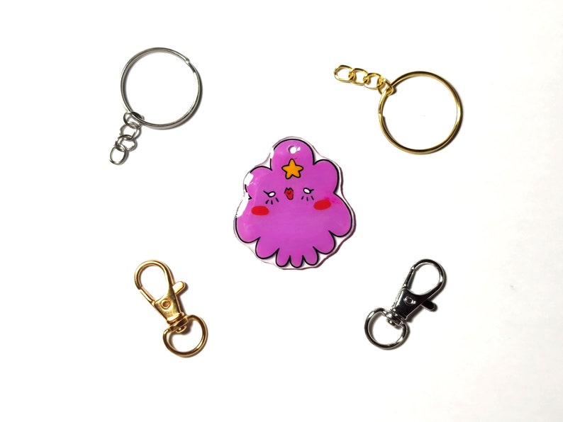 May include: A purple cartoon character keychain with a star on its head. The keychain is attached to a silver key ring. There are three other key rings in the image, two silver and one gold.