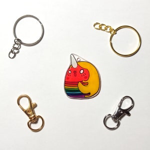 May include: A rainbow and yellow cartoon narwhal charm with a white horn, pink cheeks, and a smiling face. The charm is attached to a silver key ring with a split ring.