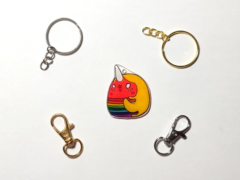 May include: A rainbow and yellow cartoon narwhal charm with a white horn, pink cheeks, and a smiling face. The charm is attached to a silver key ring with a split ring.