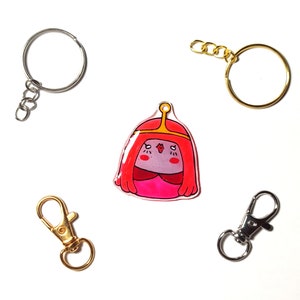 May include: A pink and red cartoon character keychain with a gold crown. The keychain is attached to a silver key ring with a chain.