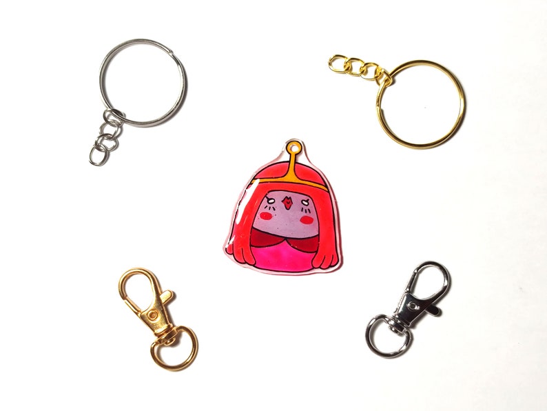 May include: A pink and red cartoon character keychain with a gold crown. The keychain is attached to a silver key ring with a chain.