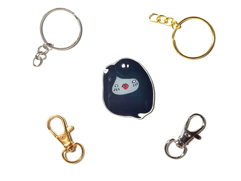 May include: Four keychains with different styles. One keychain is a silver circle with a silver chain. Another keychain is a gold circle with a gold chain. The third keychain is a silver lobster clasp. The fourth keychain is a black and white enamel pin of a cartoon character with a red mouth.