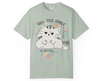 May include: A pale green t-shirt with a cartoon cat graphic and the text "Bro this kibble is still mid. Do better...". The cat is white with grey stripes and is surrounded by flowers. The shirt has a crew neck and short sleeves.