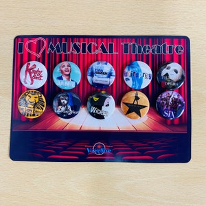 May include: A display of ten round buttons featuring various Broadway musical logos and characters. The buttons are arranged in two rows of five on a black background with a red curtain backdrop. The display is labeled "I♡MUSICAL Theatre" and "V-cresive".